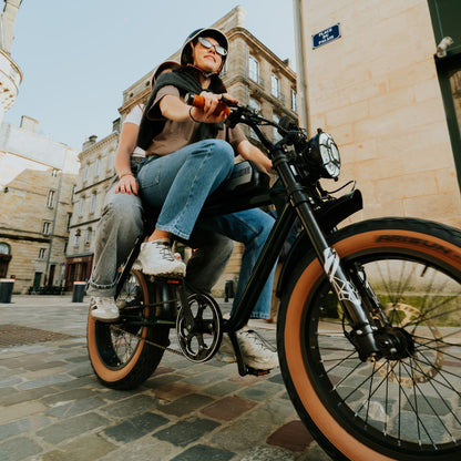 Fatbike électrique 2 places City S Unikride avec deux personnes à bord dans une rue pavée en centre-ville.